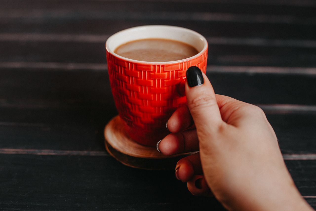 Close-up of a woman's hand with black nails holding a red coffee cup on a wooden table.